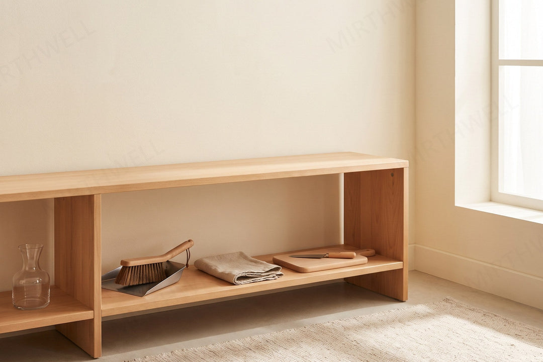 A low wooden shelf holding a small glass jug, a short-handled brush and dustpan, a folded linen cloth and a child-sized wooden board with a small knife.
