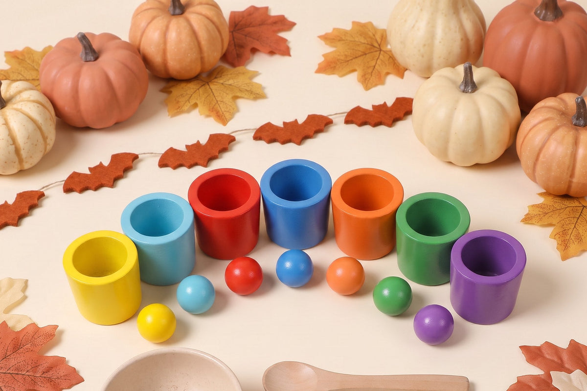 Seven wooden sorting cups in rainbow colors with a matching wooden ball in front of each, on a cream table with small pumpkins, autumn leaves and a garland of rust-orange felt bats.