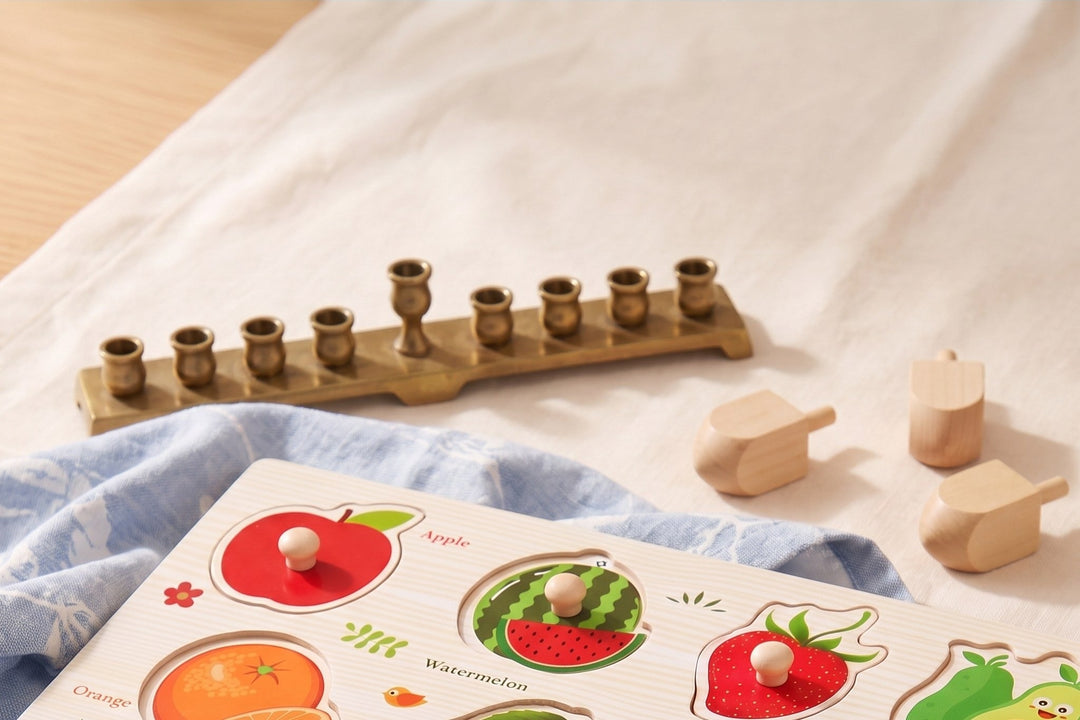 A wooden fruit peg puzzle with small wooden knobs on its pieces, on a cream linen tablecloth, with a brass Hanukkah menorah, three plain wooden dreidels and a pale blue napkin behind it.