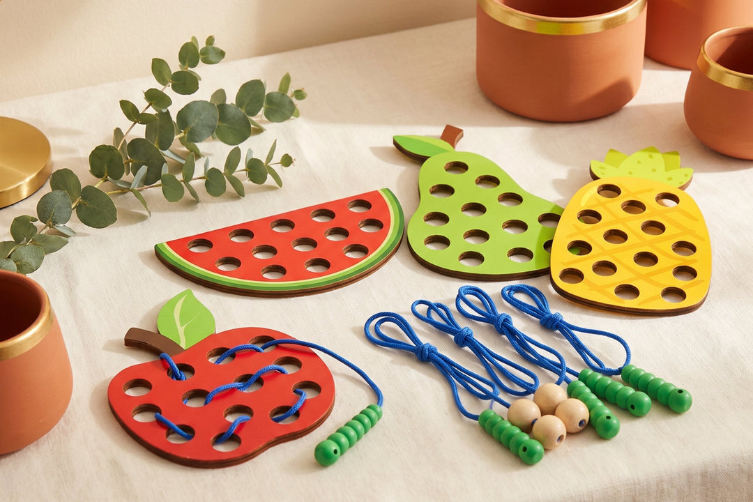 Four wooden fruit-shaped lacing boards - watermelon, apple, pear and pineapple - laid on a cream tablecloth beside blue cords with wooden worm tips and green beads, with eucalyptus and warm terracotta pots at the edges.