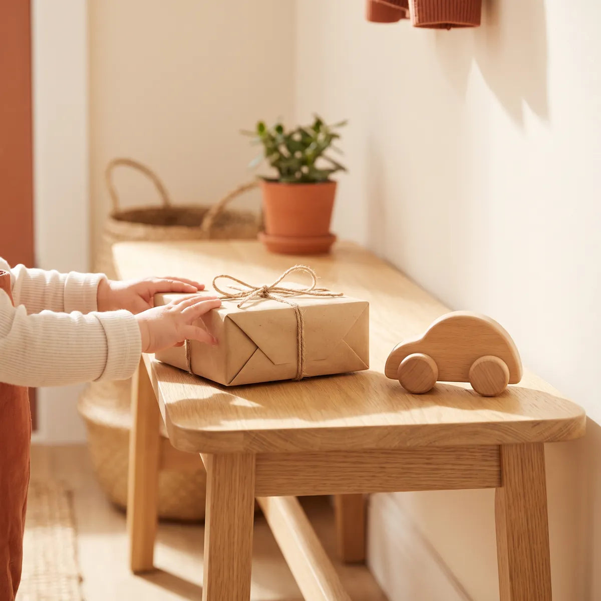 A wrapped parcel just delivered on an entryway bench, a toddler reaching for it beside a wooden toy car