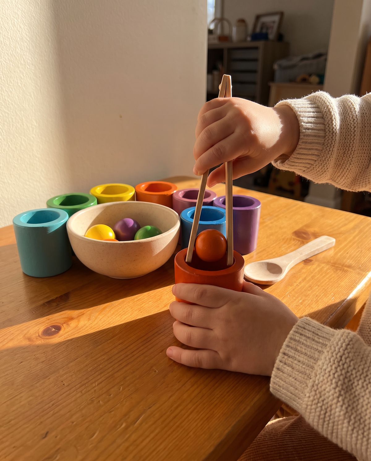 A two-year-old's hands lifting a wooden ball into a color-sorting cup at a wooden table, in afternoon light